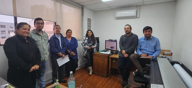 Siete profesionales posan en una oficina de la Seremi durante la capacitación en la Región de Antofagasta; al fondo, una pantalla con un visor de mapas y un plóter de gran formato.