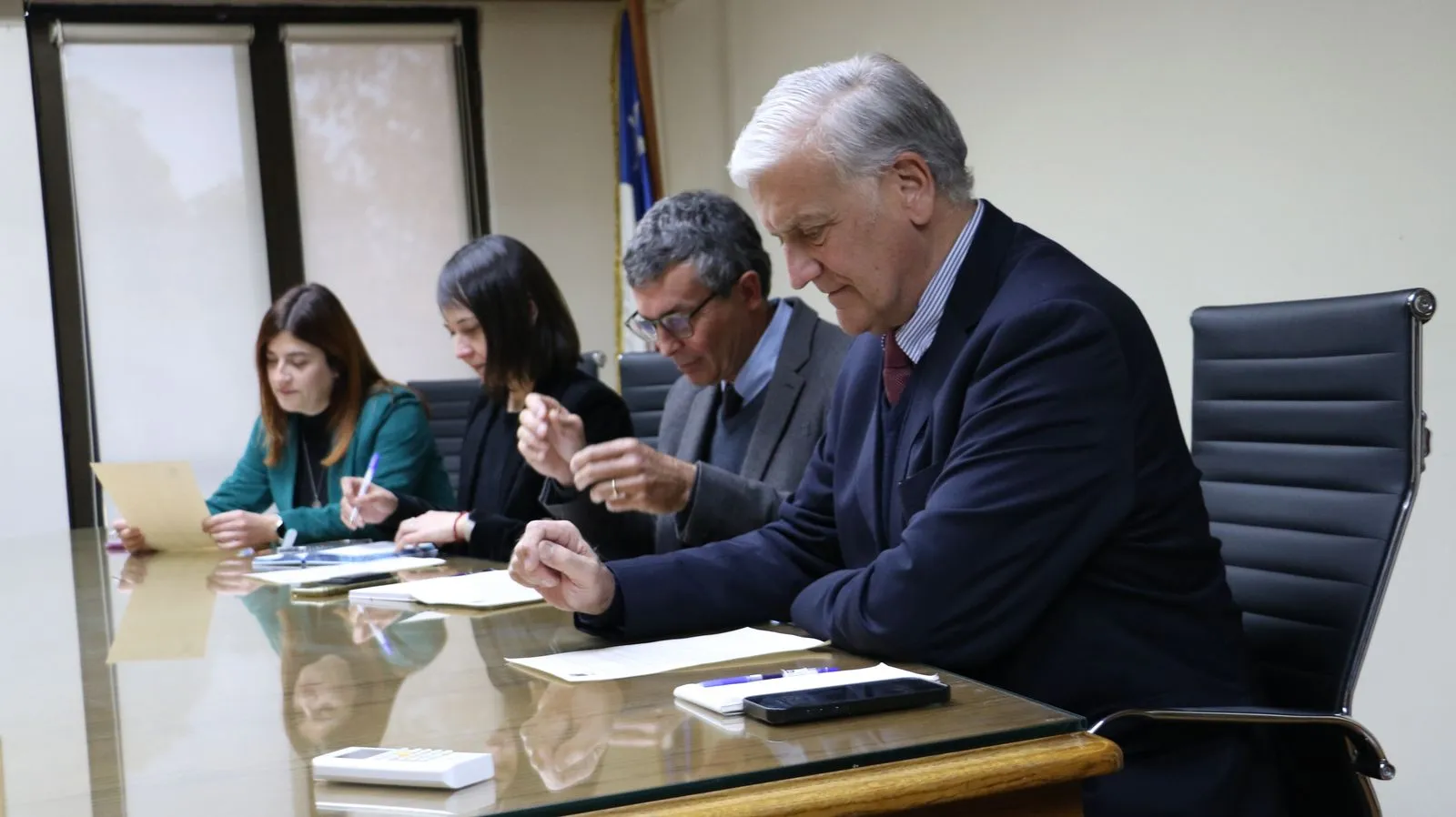 Cuatro personas firman documentos sentadas a un lado de la mesa de reuniones.