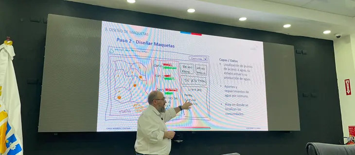 El relator señala una pantalla con la diapositiva «Paso 2 — Diseñar maquetas», que muestra un boceto de capas y datos: puntos de acceso a agua, aportes y requerimientos por comuna, y áreas donde se localizan las comunidades.
