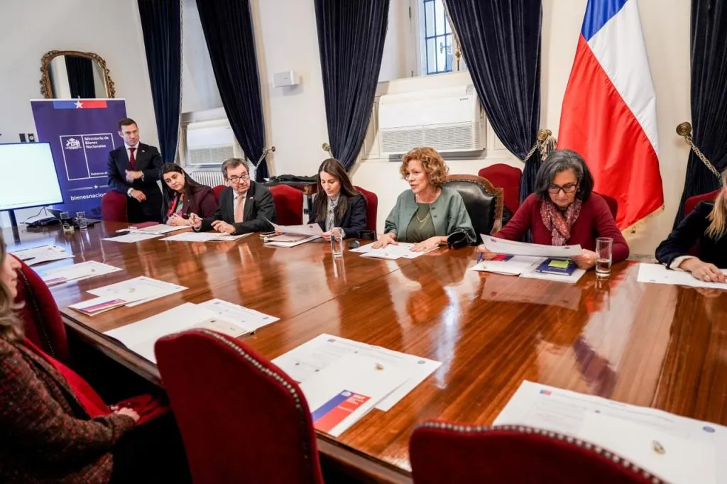Vista general de la mesa de la sesión: las autoridades revisan los documentos; al fondo, un pendón del Ministerio de Bienes Nacionales, una pantalla y la bandera de Chile.