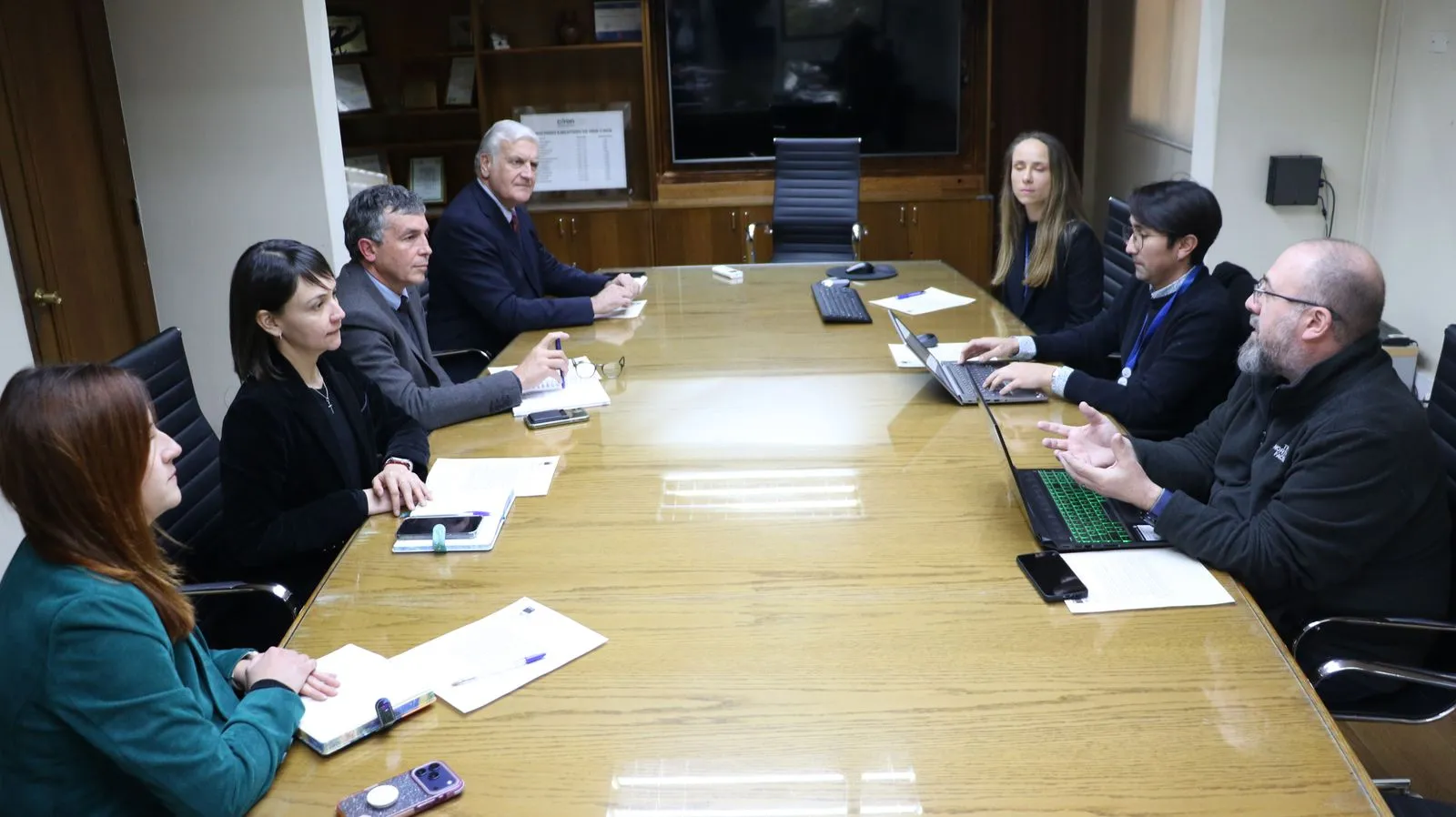 Los asistentes conversan a ambos lados de una mesa de reuniones alargada, con documentos y portátiles abiertos.