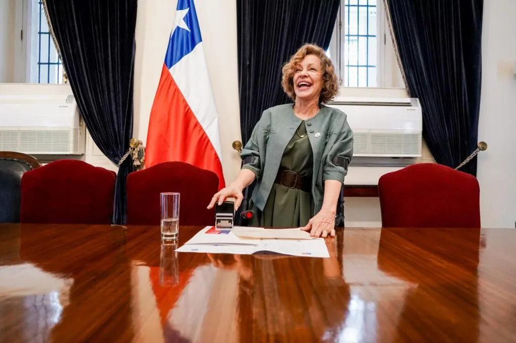 La ministra Catalina Parot, de pie tras la mesa y junto a la bandera de Chile, apoya un timbre sobre el documento de los acuerdos.