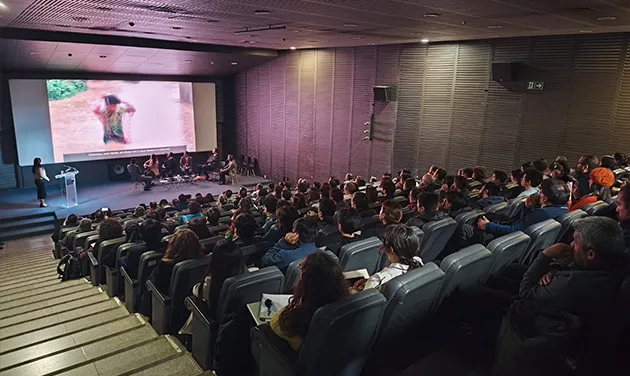 Vista de la sala llena durante una de las sesiones, con el público en las butacas y la mesa de panelistas junto a la pantalla.
