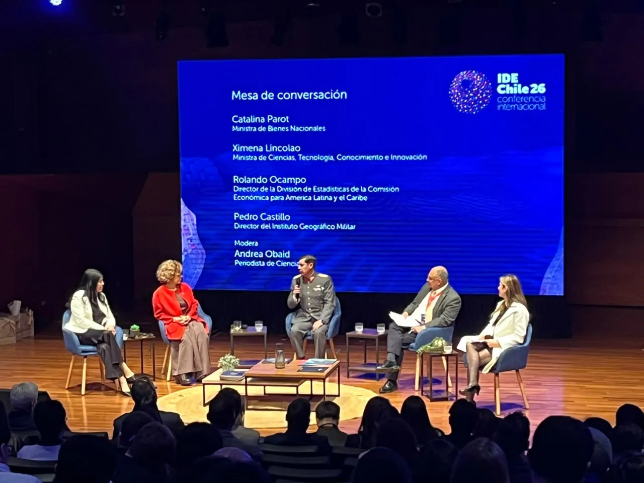 Las cuatro autoridades de la mesa y la periodista que la modera, sentadas en butacas sobre el escenario del Salón A1. Al fondo, una pantalla con el título «Mesa de conversación», sus nombres y sus cargos, junto al logotipo de la Conferencia Internacional IDE Chile 26.